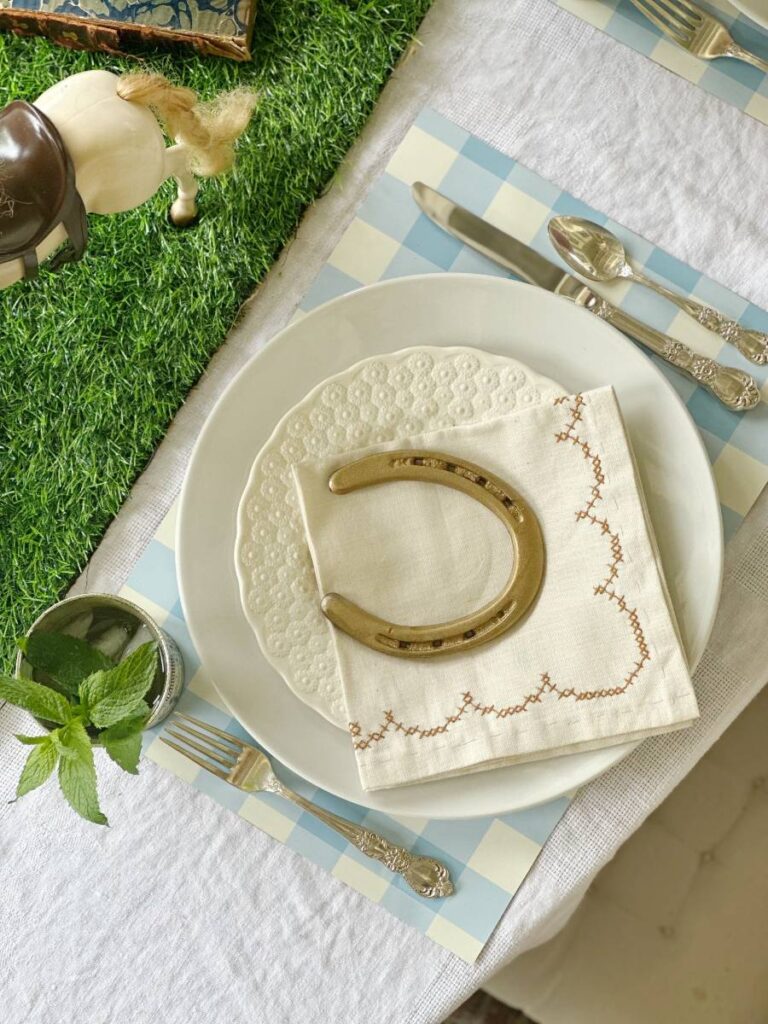 white dinner plate stacked with white salad plate folded napkin on top and gold horseshoe on blue and white check placemat and mint julep cup beside plate for Kentucky Derby place setting