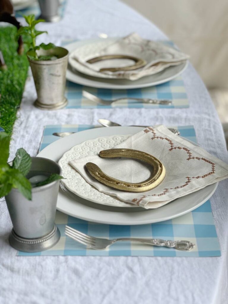 2 place settings of white dinner plate stacked with white salad plate folded napkin on top and gold horseshoe on blue and white check placemat and mint julep cup beside plate