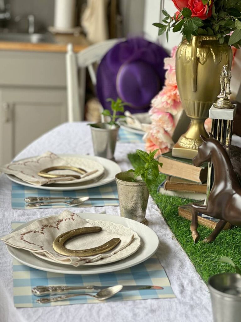 2 place settings of white dinner plate stacked with white salad plate folded napkin on top and gold horseshoe on blue and white check placemat and mint julep cup beside plate for Kentucky Derby dinner table