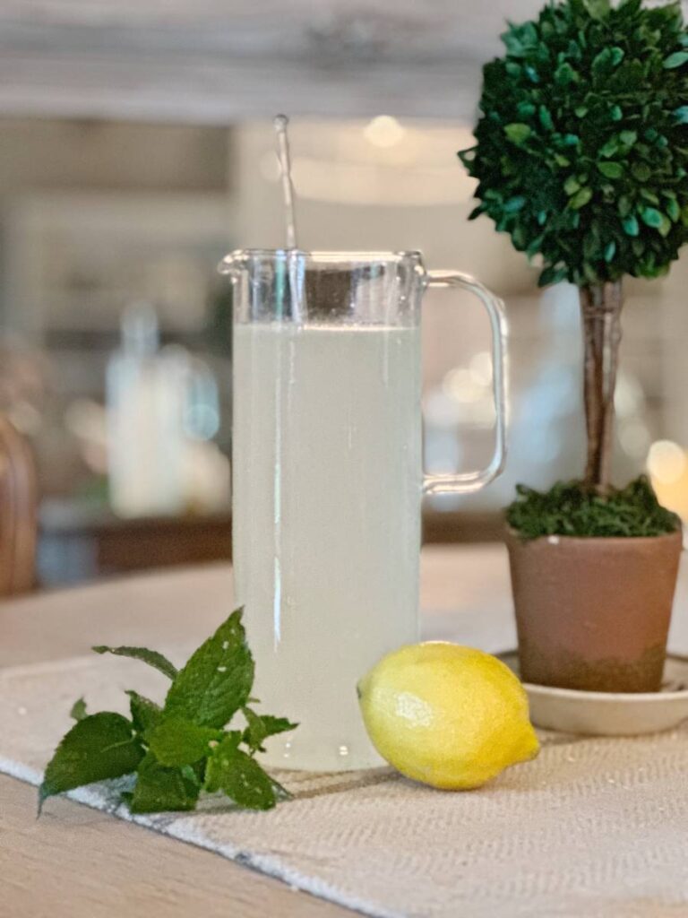 homemade lemonade in glass pitcher with lemon and mint sprig beside it