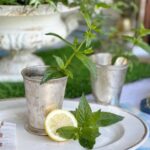 A silver cup filled with fresh mint is placed on a white plate alongside a lemon slice and more mint leaves, evoking a Mint Julep with a Lemonade Twist. In the background, another similar silver cup and a decorative green plant can be seen. The setting has a vintage and refreshing aesthetic.