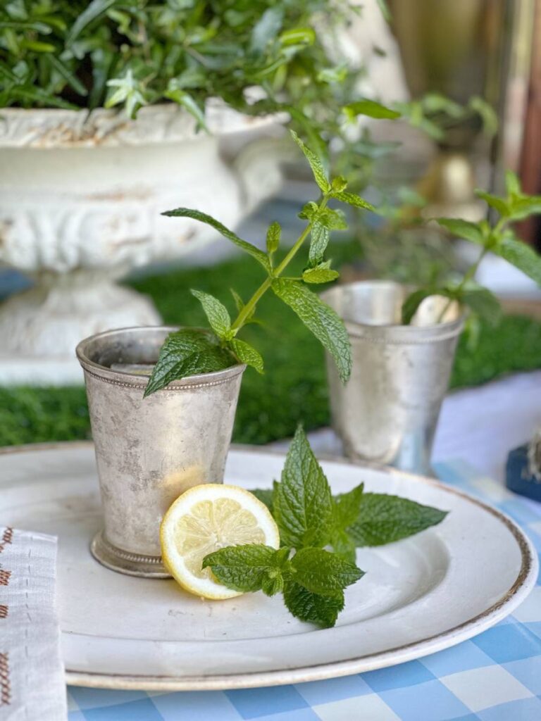 A silver cup filled with fresh mint is placed on a white plate alongside a lemon slice and more mint leaves, evoking a Mint Julep with a Lemonade Twist. In the background, another similar silver cup and a decorative green plant can be seen. The setting has a vintage and refreshing aesthetic.