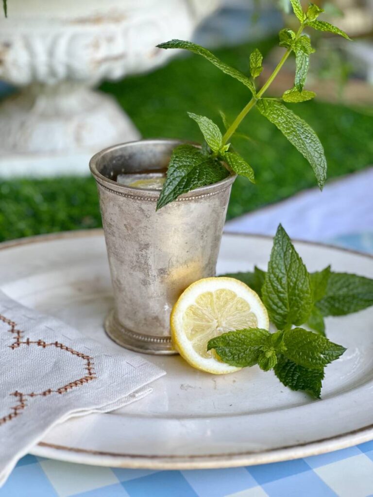 mint julep cocktail in silver julep cup with mint sprig garnish on white platter with lemon slice and mint sprig