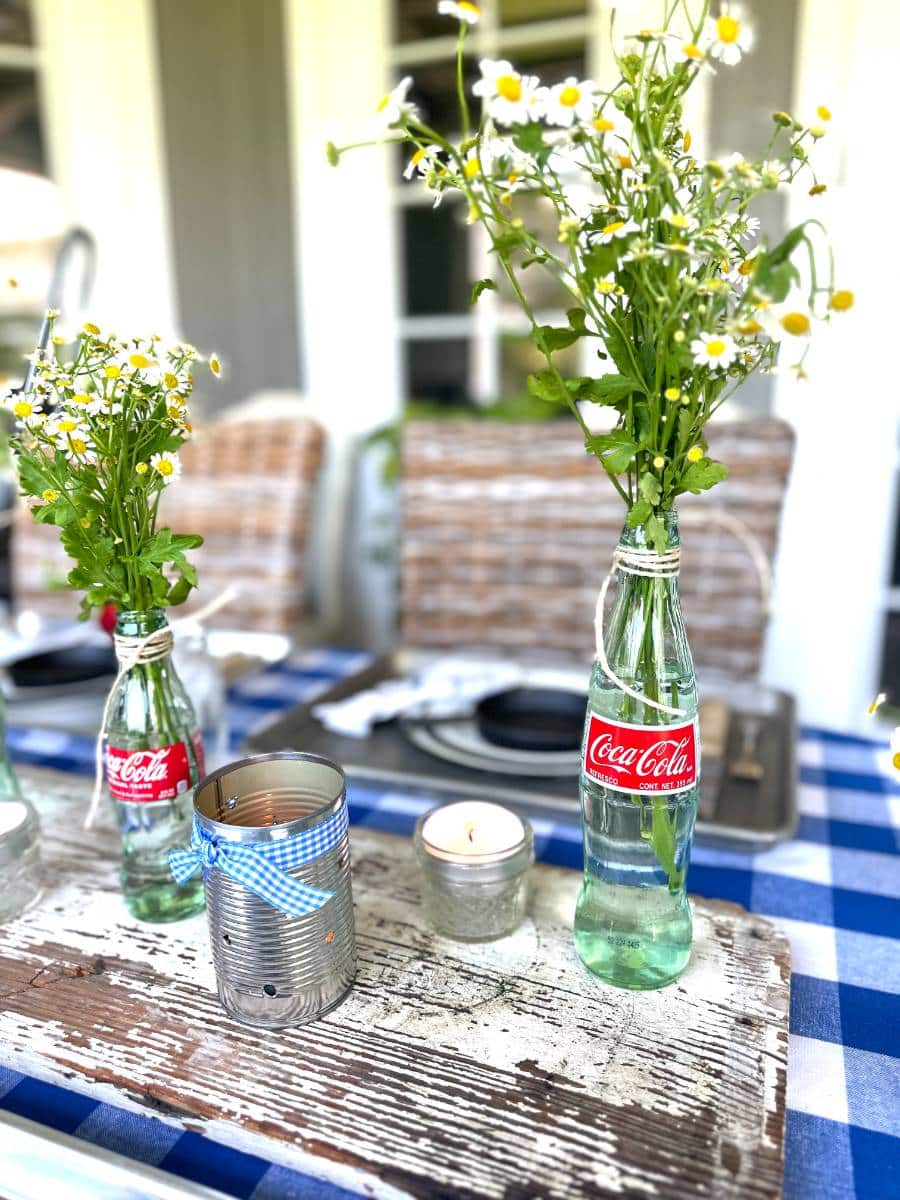white chippy board down center of table on blue and white check tablecloth 6 glass coke bottles with small white daisies tin can luminarias on top of board for table centerpiece