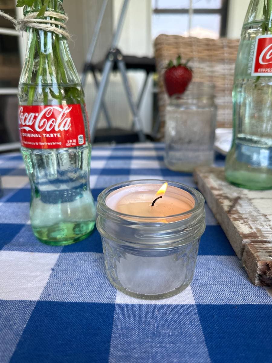 tea light candle in small mason jar beside flowers in coke bottle on blue and white check tablecloth