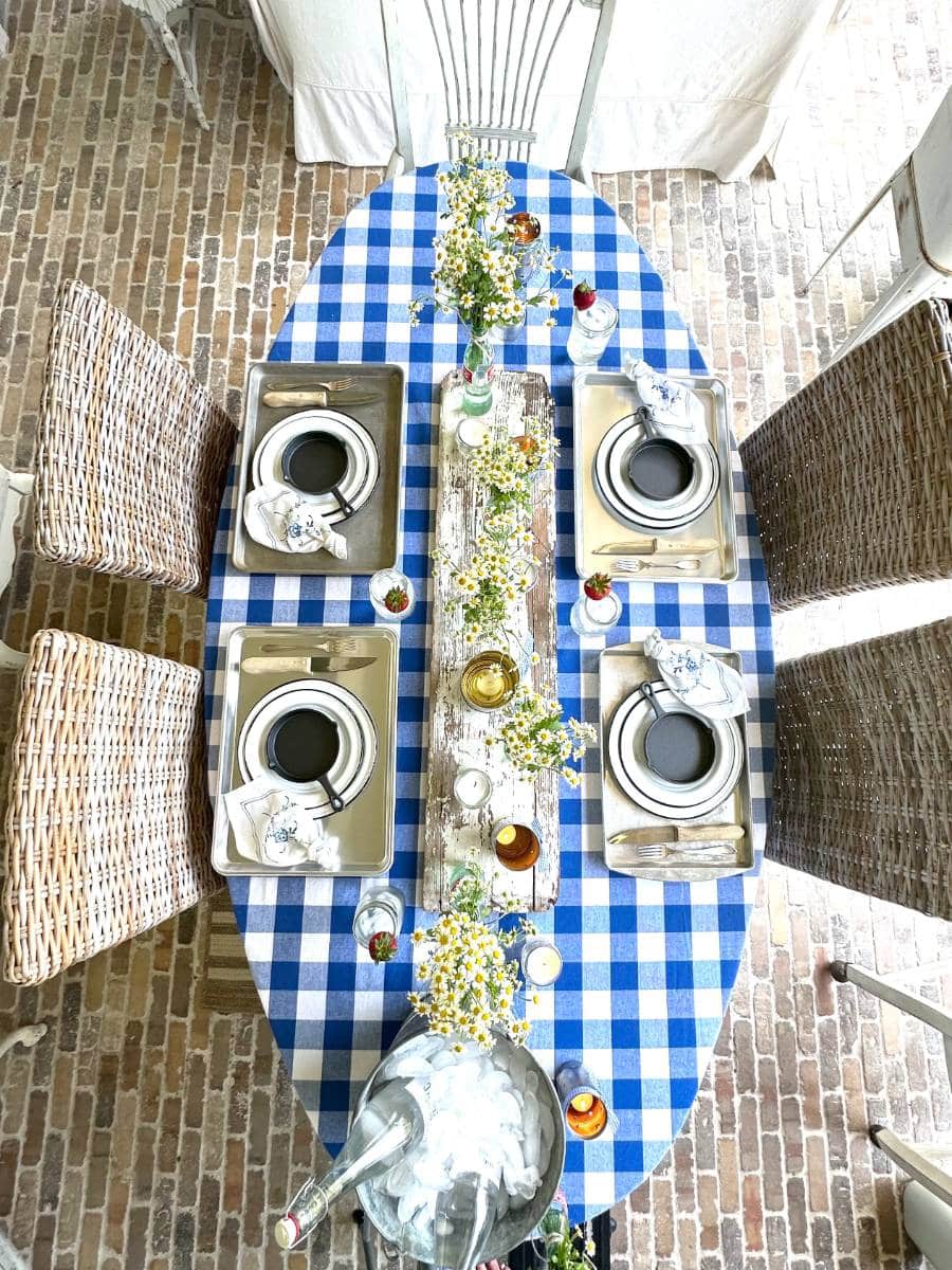blue and white check tablecloth cookie sheets at 4 place settings with layered white with black trim enamelware salad and dinner plate mini cast iron skillet on top of plates white napkin with blue floral print beside plate mason jar drinking glasses white chippy board down center of table as table runner 6 glass coke bottles with small white daisies tin can luminarias tea lights in small mason jars down center of table