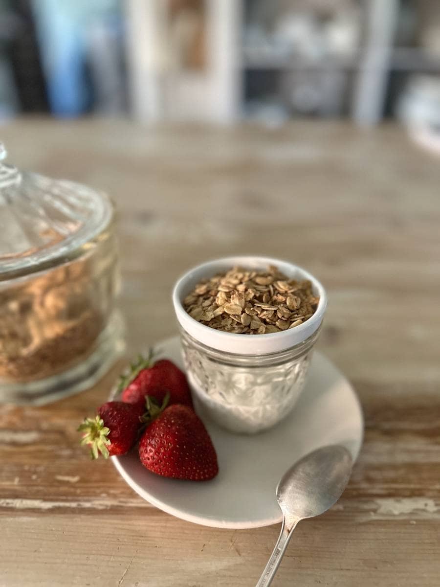 yogurt in glass jar with small bowl of granola on top and strawberries beside