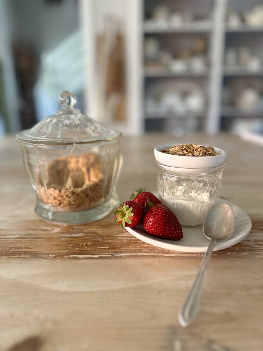 yogurt in glass jar with small bowl of granola on top and strawberries beside