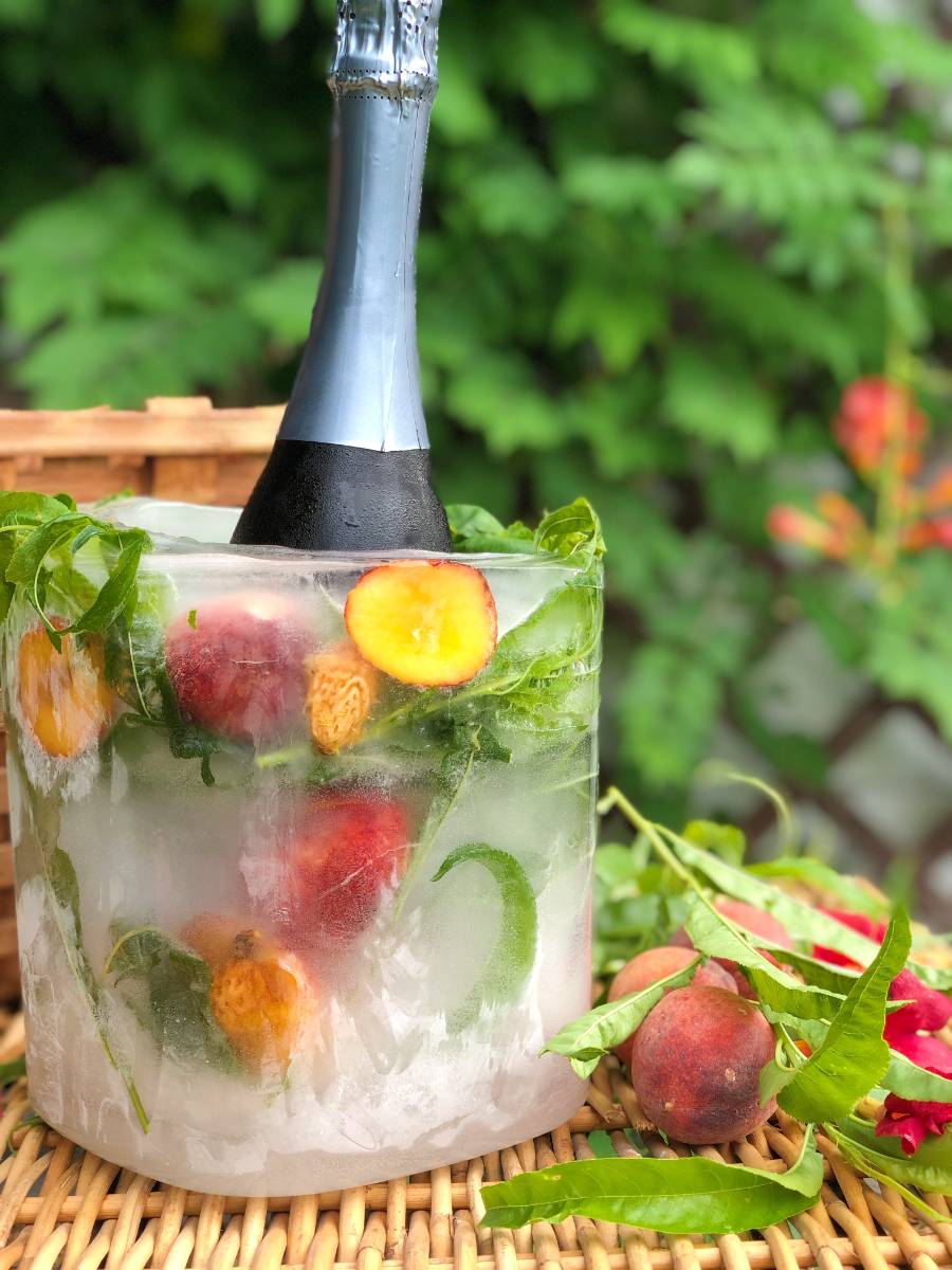 A wine bottle is encased in a cylindrical ice block containing peaches and green leaves. The ice block is set on a woven surface with green foliage in the background. A few peaches and leaves are placed around the base, enhancing the summer ambiance.