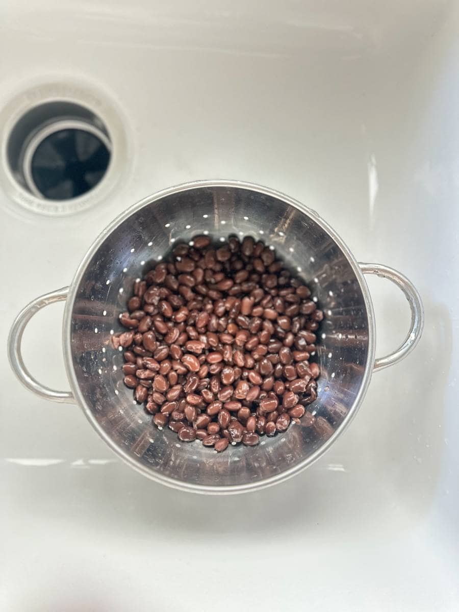 A stainless steel colander filled with rinsed black beans sits in a white sink. The sink’s drain is visible in the upper left corner. The beans appear wet and shiny, ready to be combined with corn for delicious Black Bean and Corn Nachos with Balsamic Glaze.