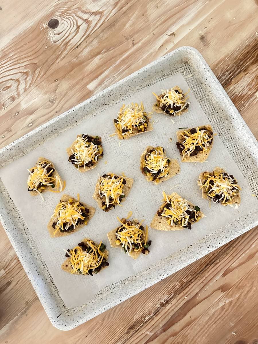 A baking tray lined with parchment paper holds ten small nacho chips, each topped with black beans, a sprinkle of corn, jalapeño slices, and a generous sprinkling of shredded cheese. These Black Bean and Corn Nachos with Balsamic Glaze are ready to be baked. The tray is set on a wooden surface.