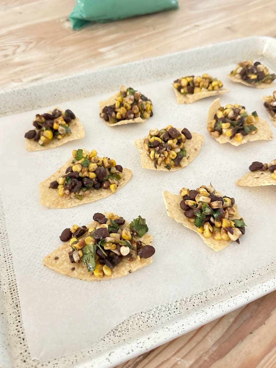 A tray of Black Bean and Corn Nachos with Balsamic Glaze is topped with a mix of black beans, corn, chopped cilantro, and other veggies on parchment paper. The tray sits on a light wooden surface, with a green bag in the background.