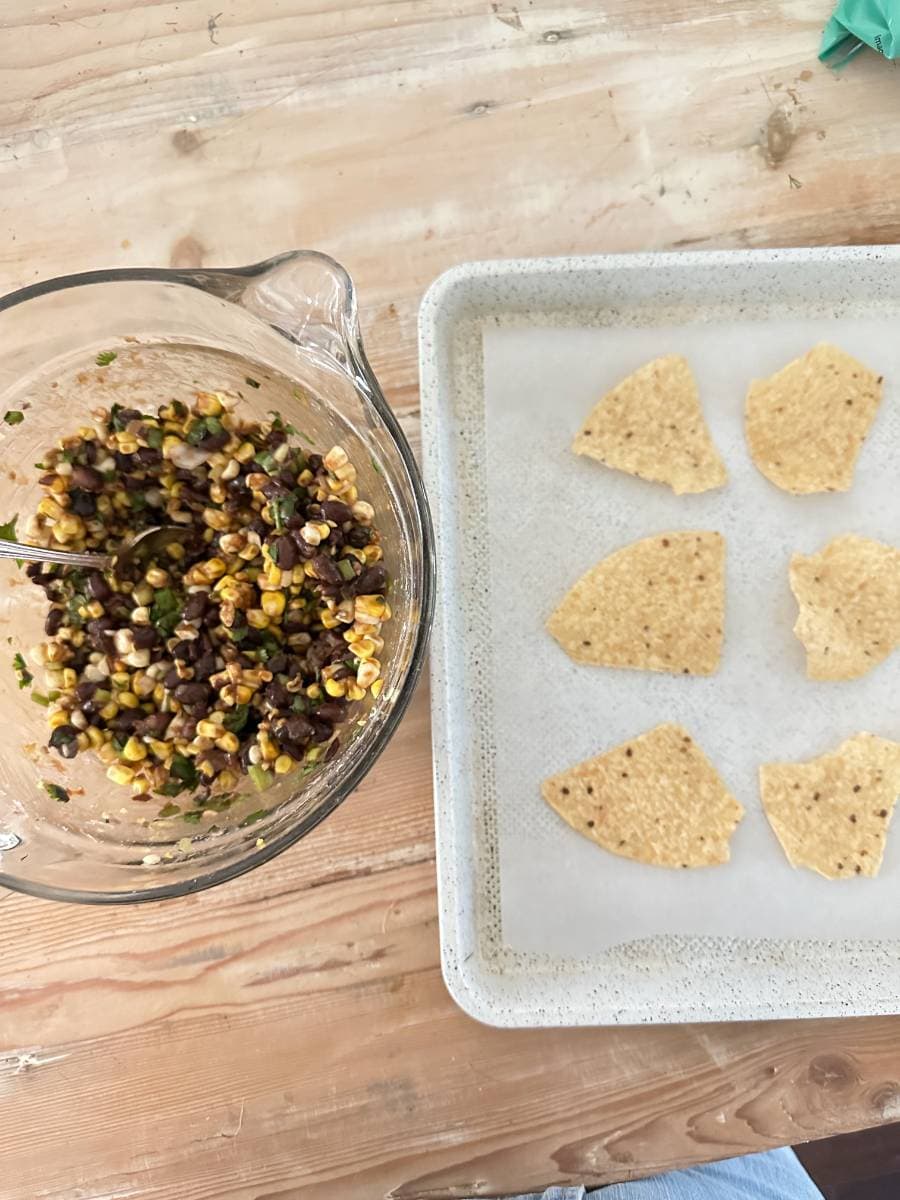 A glass bowl filled with a delicious mixture of corn, black beans, chopped vegetables, and herbs sits on a wooden table. Next to it is a baking tray lined with parchment paper holding five tortilla chip halves. A spoon is in the bowl, ready for serving Black Bean and Corn Nachos with Balsamic Glaze.