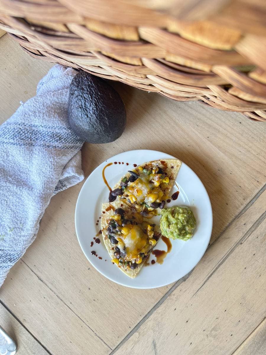 A plate of Black Bean and Corn Nachos with Balsamic Glaze, topped with melted cheese and accompanied by a dollop of guacamole on the side. The plate is set on a light wooden surface near an avocado, a wicker basket, and a white cloth.