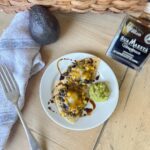 A small white plate holds Black Bean and Corn Nachos with melted cheese, garnished with a drizzle of balsamic glaze. A dollop of guacamole is also on the plate. An avocado, a fork, a towel, and a bottle of "Sua Maesta" balsamic vinegar are beside the plate.