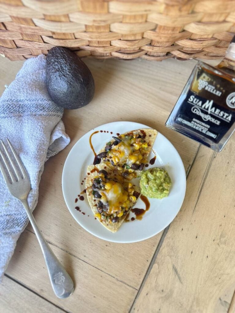 A small white plate holds Black Bean and Corn Nachos with melted cheese, garnished with a drizzle of balsamic glaze. A dollop of guacamole is also on the plate. An avocado, a fork, a towel, and a bottle of "Sua Maesta" balsamic vinegar are beside the plate.