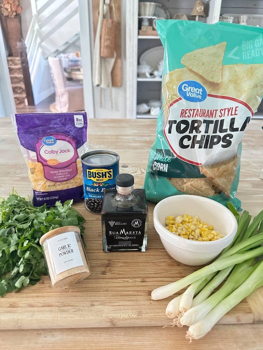 A wooden table holds ingredients for Black Bean and Corn Nachos with Balsamic Glaze: a package of Colby Jack cheese, a can of black beans, a bag of tortilla chips, fresh cilantro, garlic powder, sea salt, corn in a bowl, and fresh green onions. A kitchen is visible in the background.