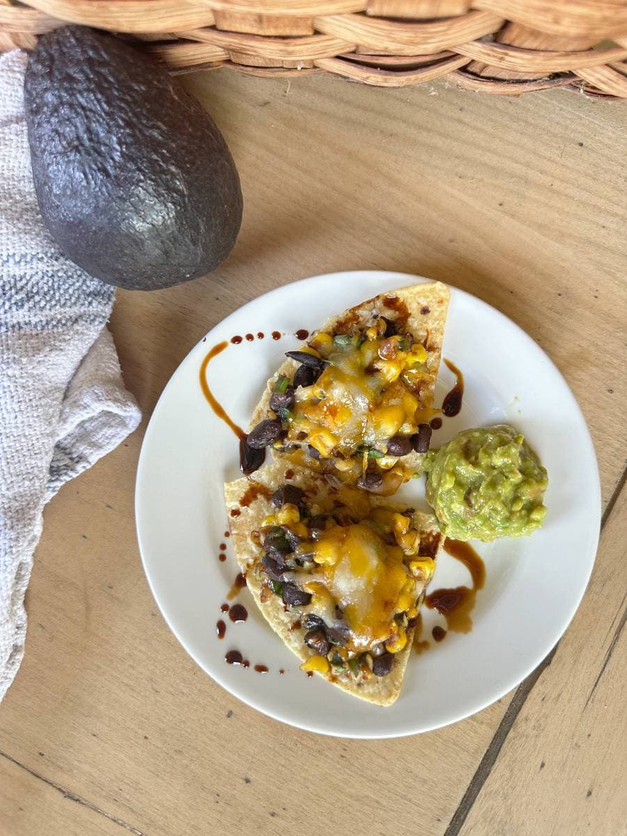 A white plate on a wooden table holds two Black Bean and Corn Nachos with melted cheese, herbs, and a delicate balsamic glaze. A scoop of guacamole sits beside them. An avocado and a woven basket are in the background.