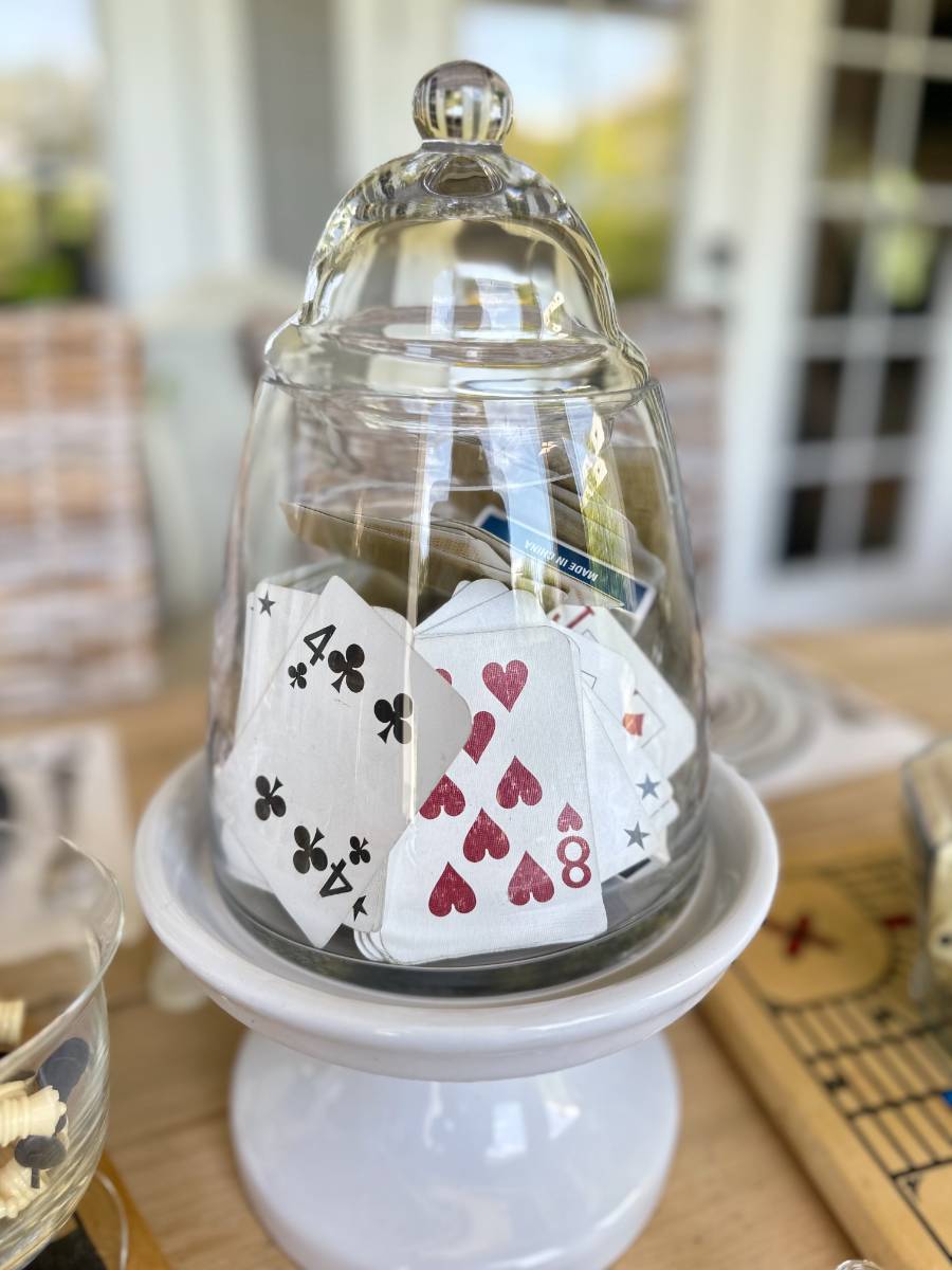 A glass cloche with a knob handle covering a cluster of playing cards, including the eight of hearts and seven of clubs, placed on a white pedestal. The background features a blurred indoor setting with books and other table items.