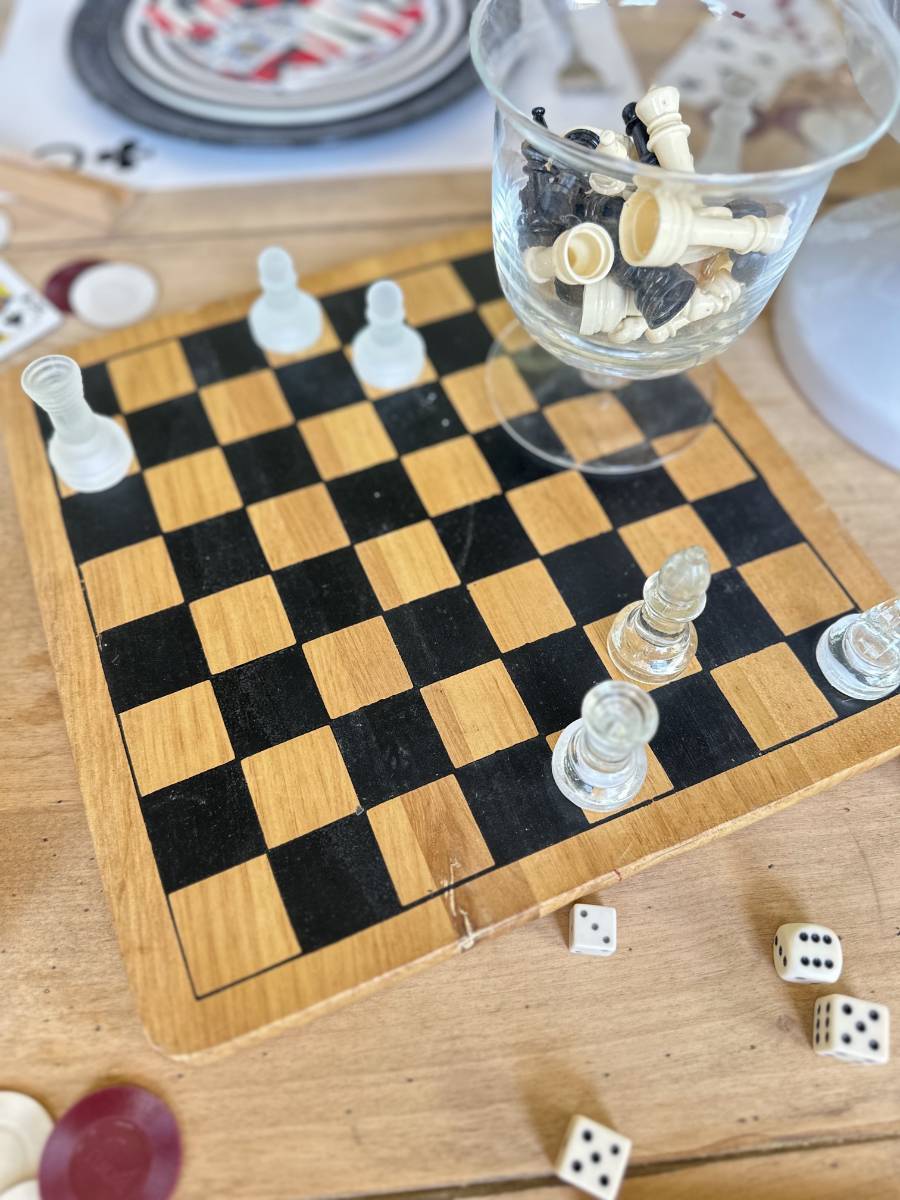 Close-up of a chessboard with four glass pieces placed on it. A clear glass is filled with various chess pieces and set on the board. Dice and poker chips are scattered around the board. The setup appears to be on a table.