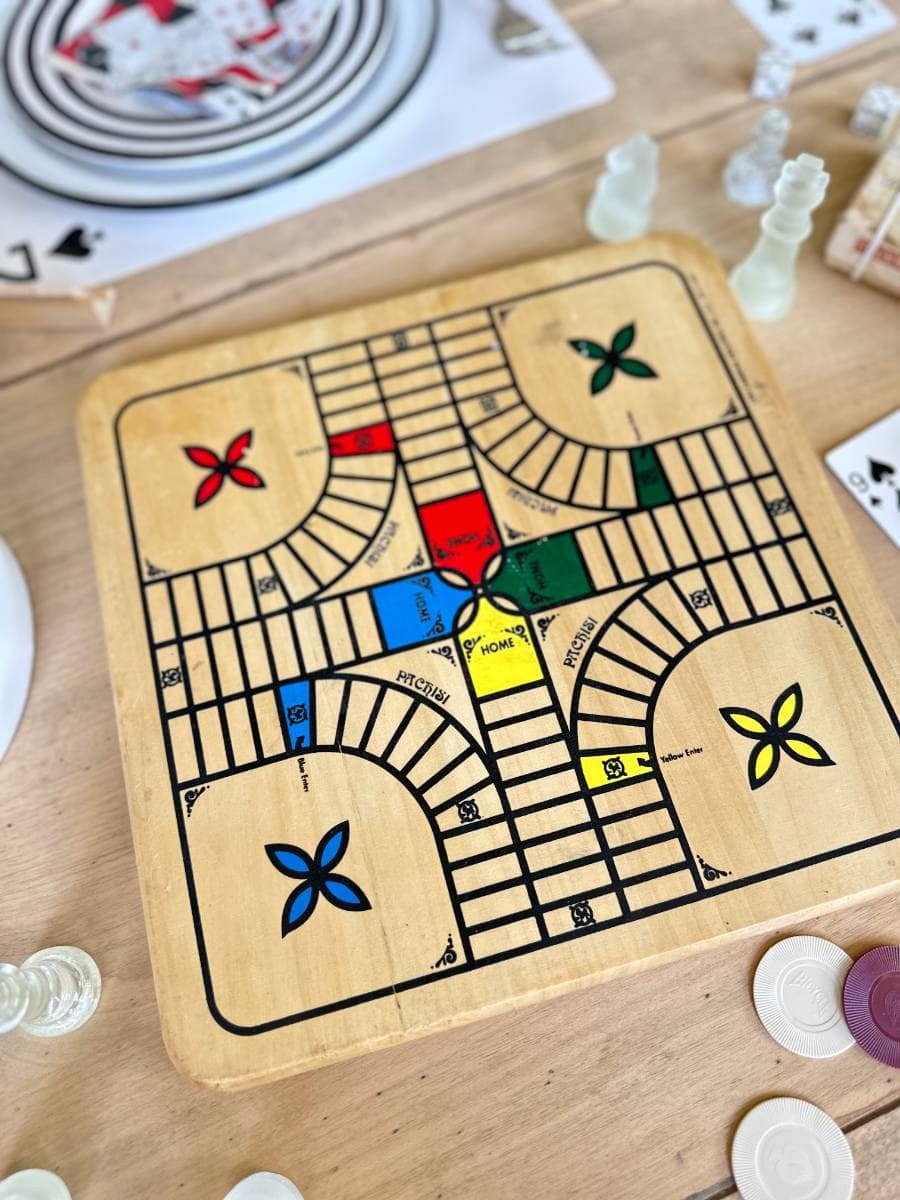 A Parcheesi board game set up on a wooden table. The board features designated home areas in red, green, yellow, and blue, with pathways leading to a central safe area. Chess pieces and chips are scattered around the table, indicating a mixed gameplay session.