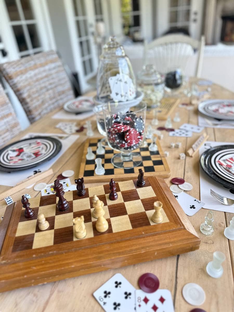 A wooden table is set up for a game night with chessboards, poker chips in glass jars, and scattered playing cards. Plates with dice-themed designs are placed at each seat. The background features wicker chairs and a partially visible doorway.