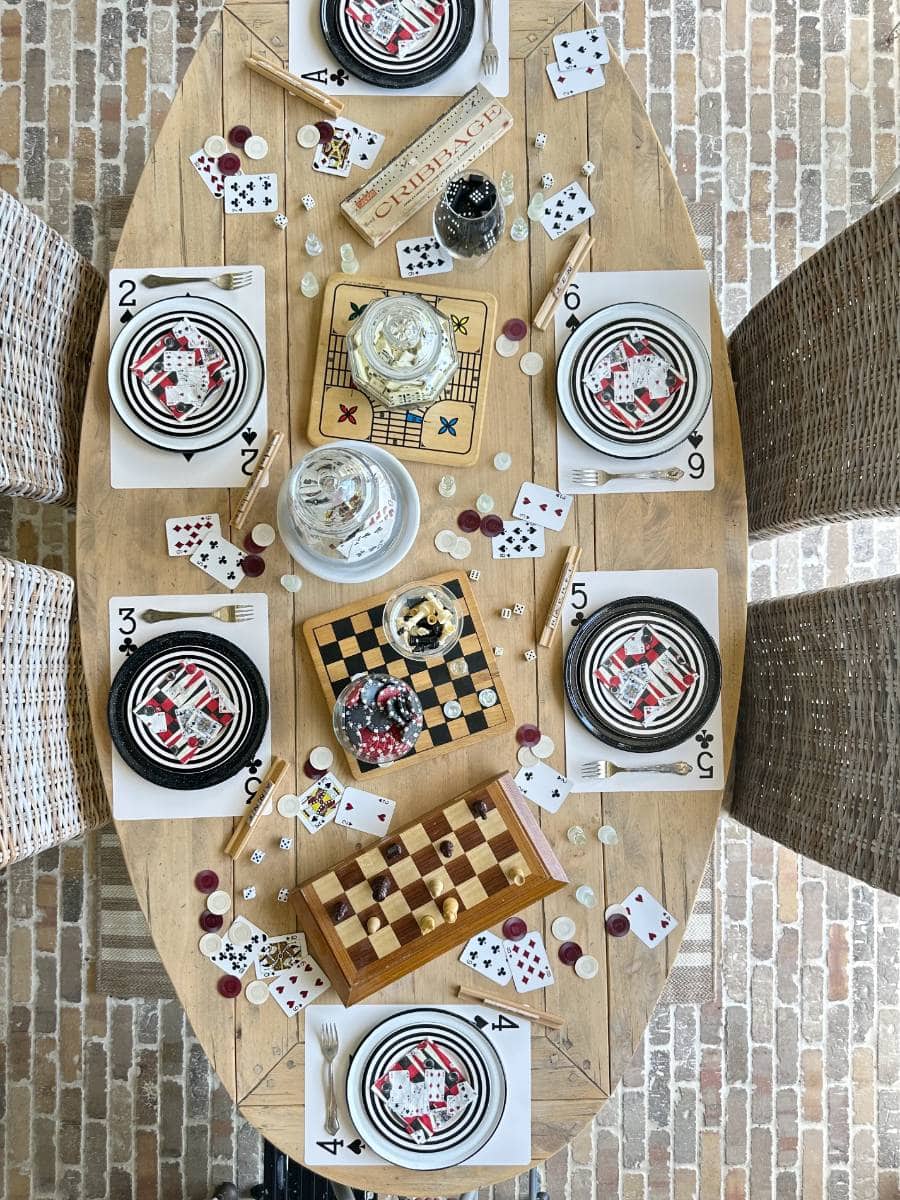 A rectangular table set for a meal is decorated with playing cards, poker chips, and black and white striped plates. A chessboard is placed at the center along with bowls of mixed nuts. Wooden chairs surround the table, and the setting is on a brick floor.