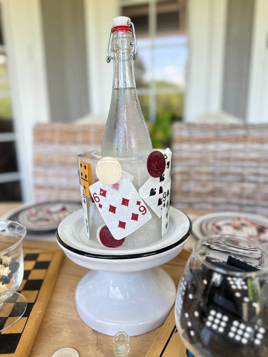 A glass bottle with a flip-top seal sits on a white pedestal plate. Several playing cards, poker chips, and a yellow die are arranged around its base. The background features wicker furniture and a window, with a blurred view of greenery outside.