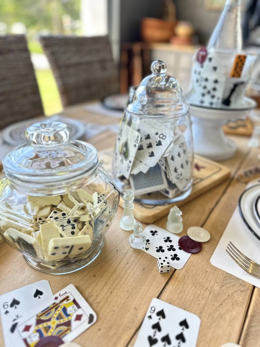 A wooden table displays two glass jars, one filled with dominoes and the other with playing cards. Scattered cards, poker chips, and dice adorn the table. Place settings with white plates are visible in the background, and wicker chairs surround the setup.