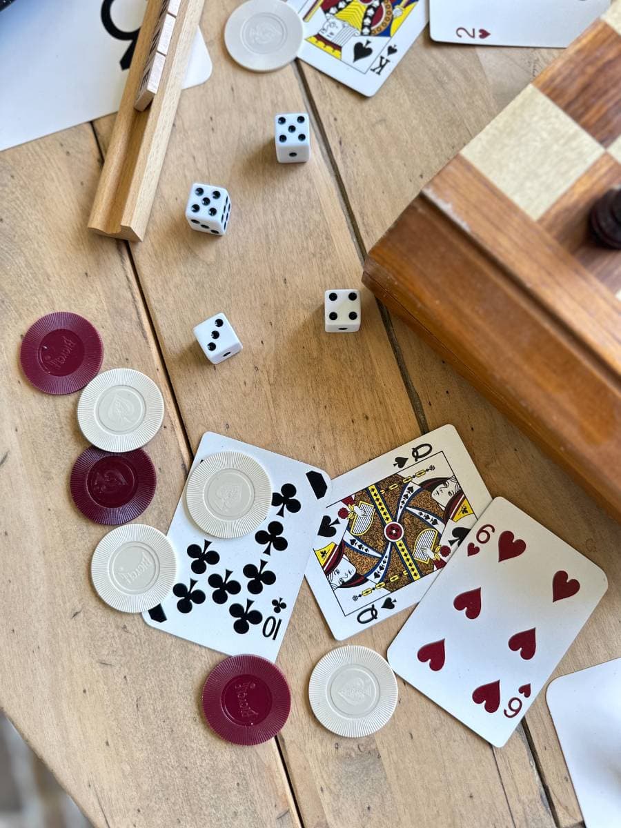 A wooden table is scattered with playing cards, poker chips in red and white, four six-sided dice, and wooden dominoes. Cards visible include the 10 of clubs, the King of diamonds, and the 6 of hearts. An edged wooden box sits at the top right corner of the table.