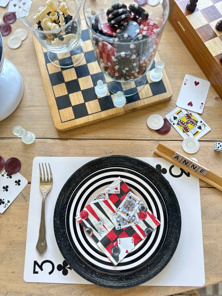 A tabletop scene features a plate with black and white spiral patterns, holding a folded cloth with card suits design. Around it are chess pieces, poker chips, playing cards, and a Scrabble tile holder displaying the name "ANNE." A fork is laid on a placemat with a club suit design.