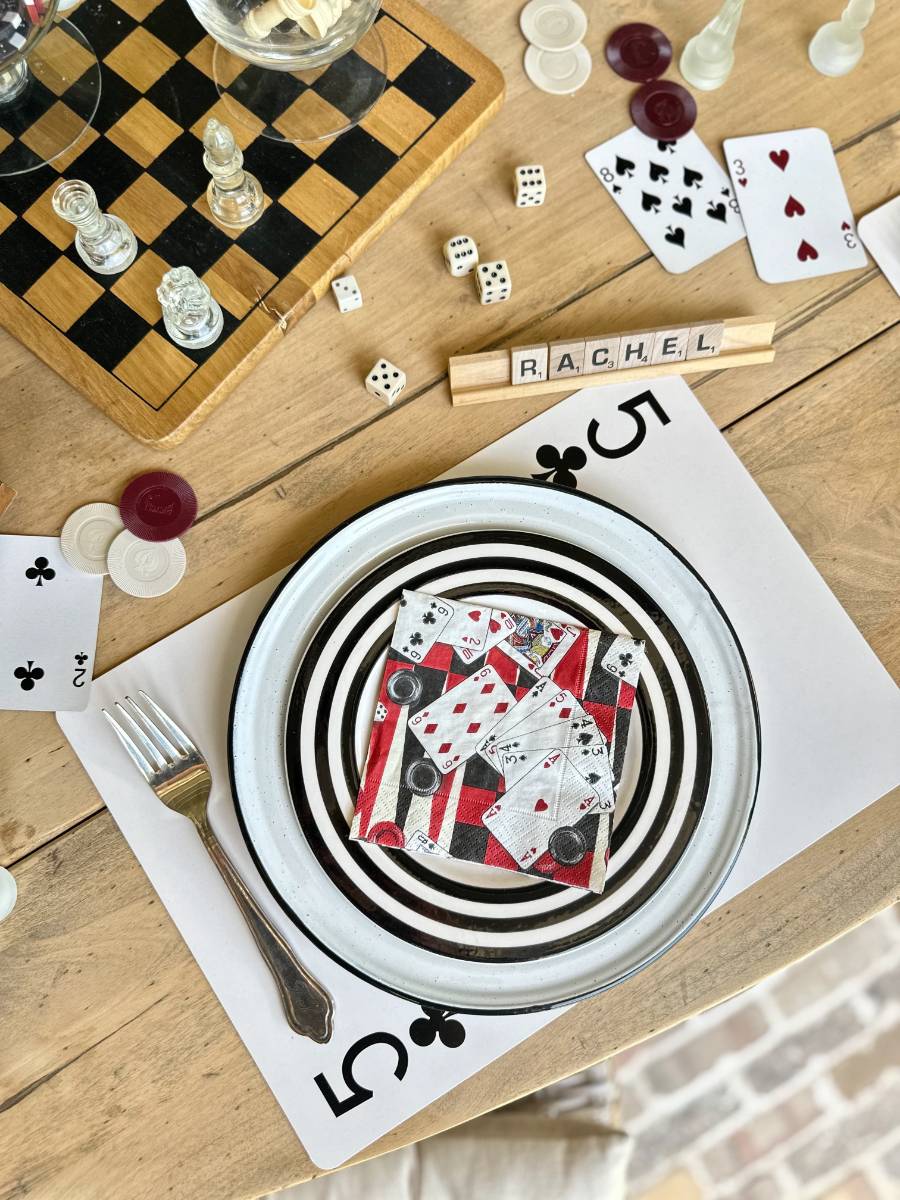 A cluttered table displays a mix of games and dinnerware: a chessboard with pieces, playing cards, Scrabble tiles spelling "RACHEL," dice, poker chips, and a fork. A plate with a swirl design holds scattered playing cards, all atop a large 5 of clubs card.