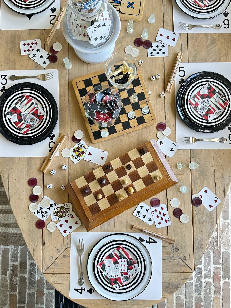 A wooden table is set for four with plates on playing card-themed placemats and napkins featuring red and black designs. The table is decorated with dice, poker chips, chess pieces, and playing cards. A tiered stand and a checkerboard holding candies and beads are at the center.