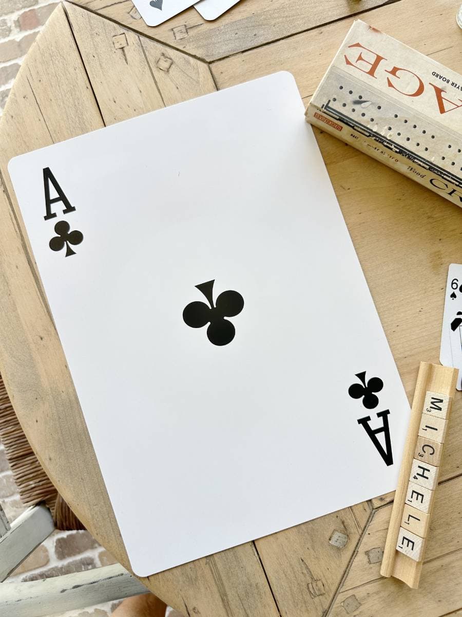 A large ace of clubs card is placed on a wooden table. Nearby, there's a box marked "ACE," a set of dominoes, and a rack of Scrabble tiles spelling "MICHELE." A few other cards are partially visible in the background.