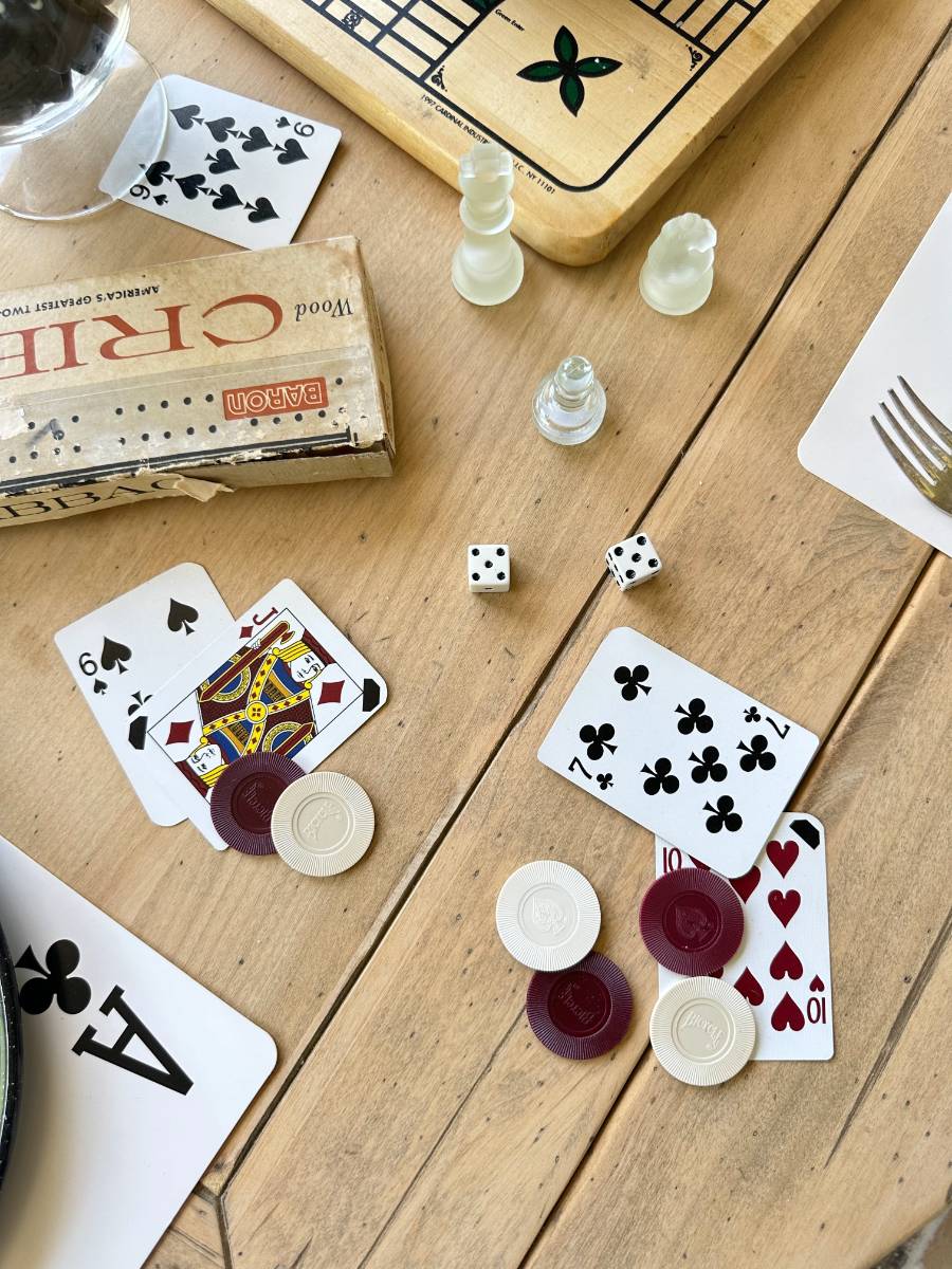 A casual game scene on a wooden table with playing cards, poker chips, two dice, a board game in the background, and a partial visible dinner setting with a fork and knife. The focus is on the cards and chips, suggesting a friendly game night.