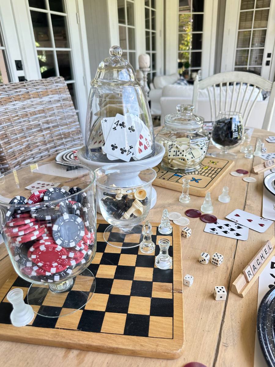 A wooden table set for a game night is adorned with various game pieces and decorations. There are jars filled with poker chips, playing cards, dice, and chess pieces. A Scrabble stand spells out "RACHEL". Board games and playing cards are spread across the table.