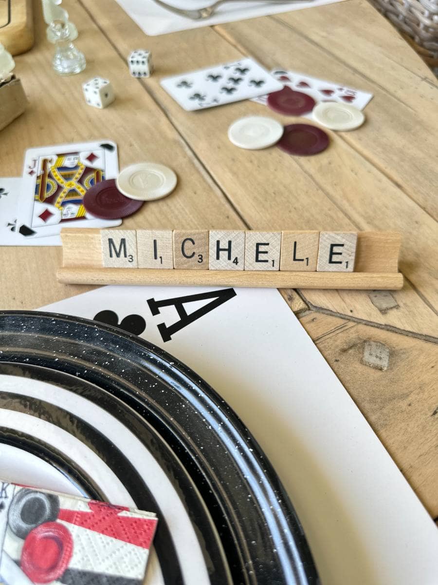 A wooden Scrabble letter holder spells out "MICHELE" with Scrabble tiles. Surrounding items include playing cards, poker chips, dice, a striped plate, and scattered gaming paraphernalia on a wooden table.