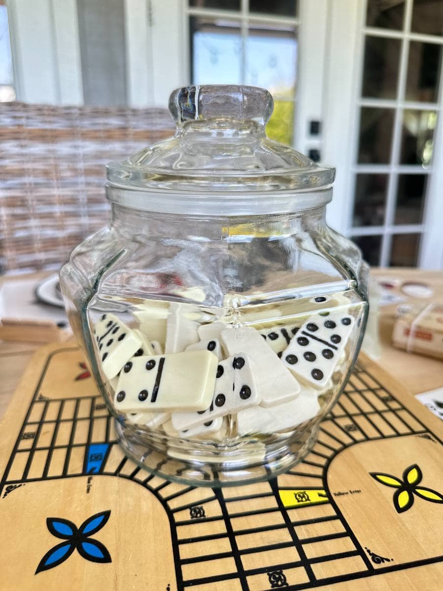 A clear glass jar filled with white dominoes featuring black dots sits on a Ludo game board. The scene is set indoors, with a wicker chair and glass-paneled doors visible in the background.