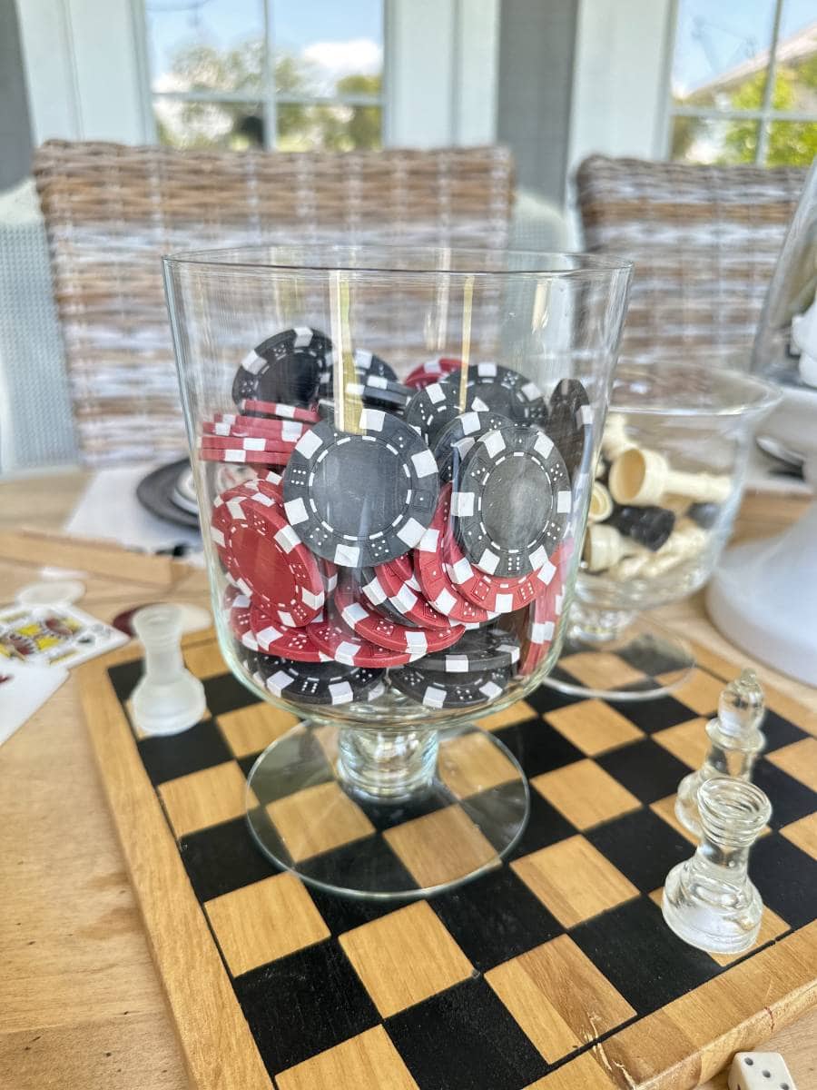 A glass container filled with poker chips in red, black, and white is placed on a table. In the foreground, there is a chessboard with a few chess pieces, including a black king and white pawn. White wicker chairs and a window are visible in the background.