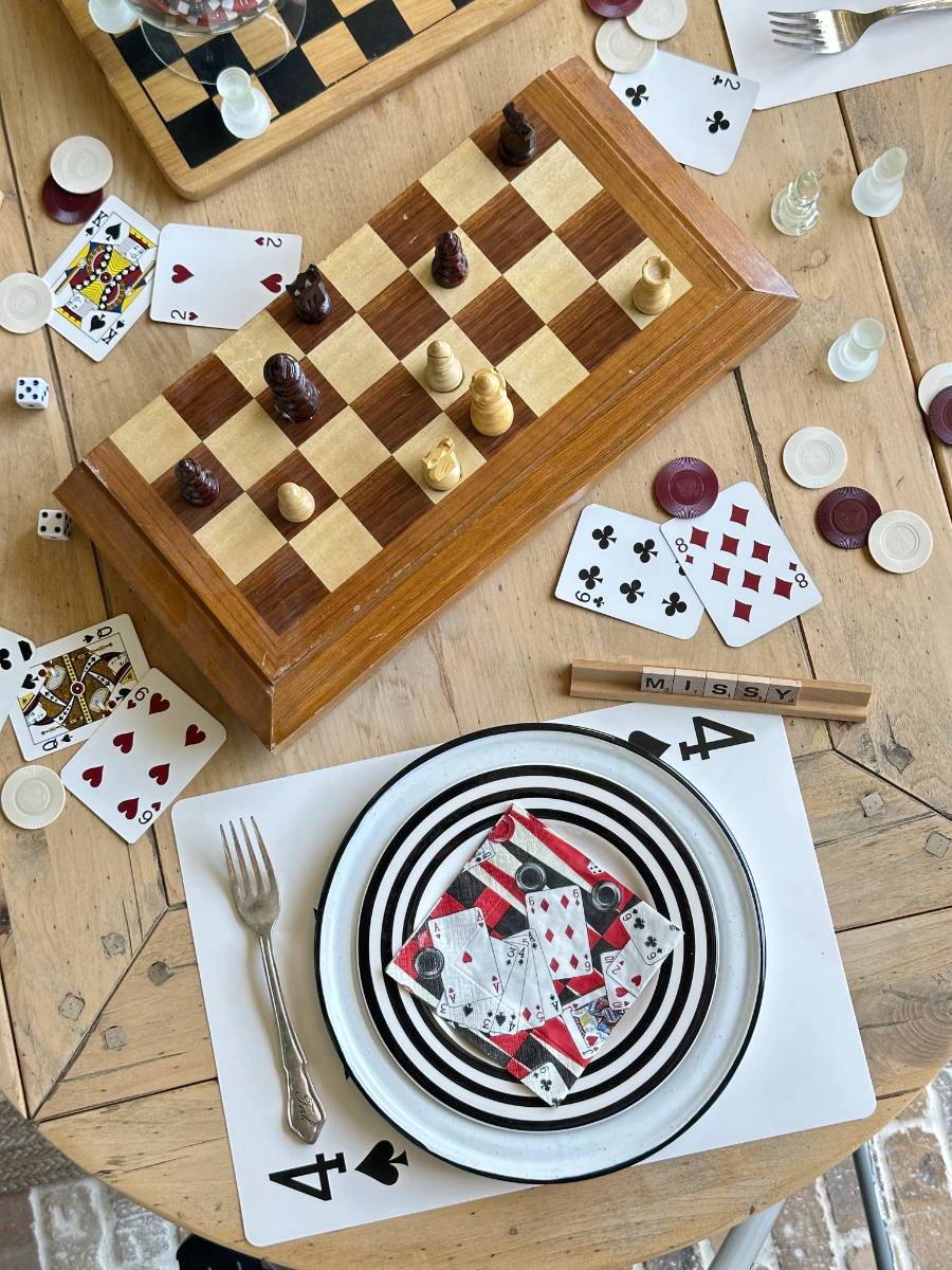 A wooden table is strewn with playing cards, poker chips, a checkered chessboard with chess pieces, a fork, and a plate holding a ring of playing cards. Dice, a small notepad with pens, and placemats featuring the Ace of Spades are also seen.