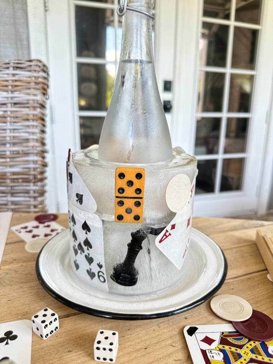 A chilled bottle is encased in a block of ice adorned with playing cards, poker chips, a domino, a chess piece, and a die, all set on a plate. Surrounding the plate are additional cards, dice, and poker chips. A door with glass panels is in the background.