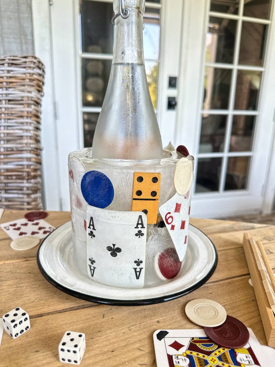 A frosted bottle of wine is chilled in a decorative ice bucket covered with playing cards and poker chips. The scene is set on a rustic table with a glass door and wicker chair in the background. Dice and additional playing cards are scattered around the table.