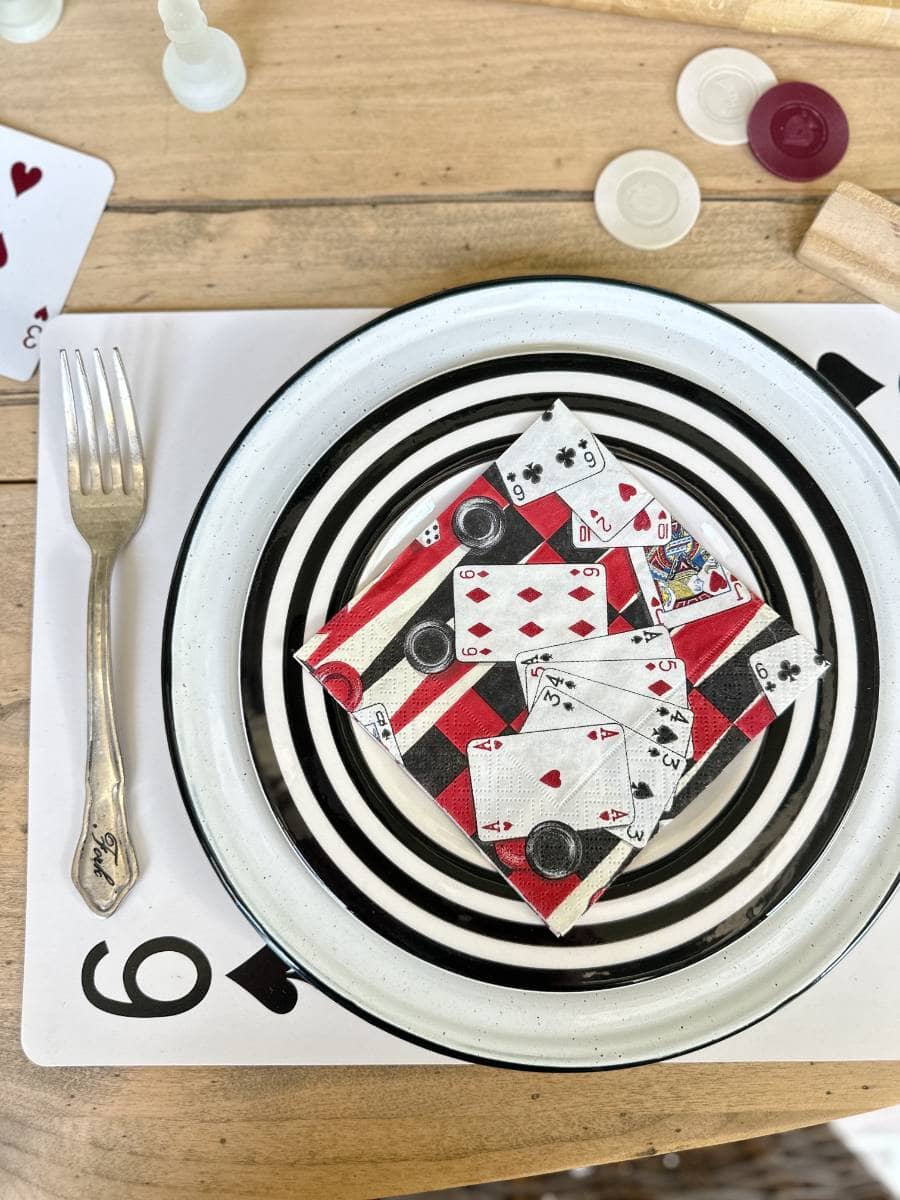 A plate on a table is decorated with playing card napkins folded into squares and topped with game chips. Next to the plate is a fork, and under it is a placemat featuring a large number '9' and a spade symbol. The table also has various game pieces scattered around.