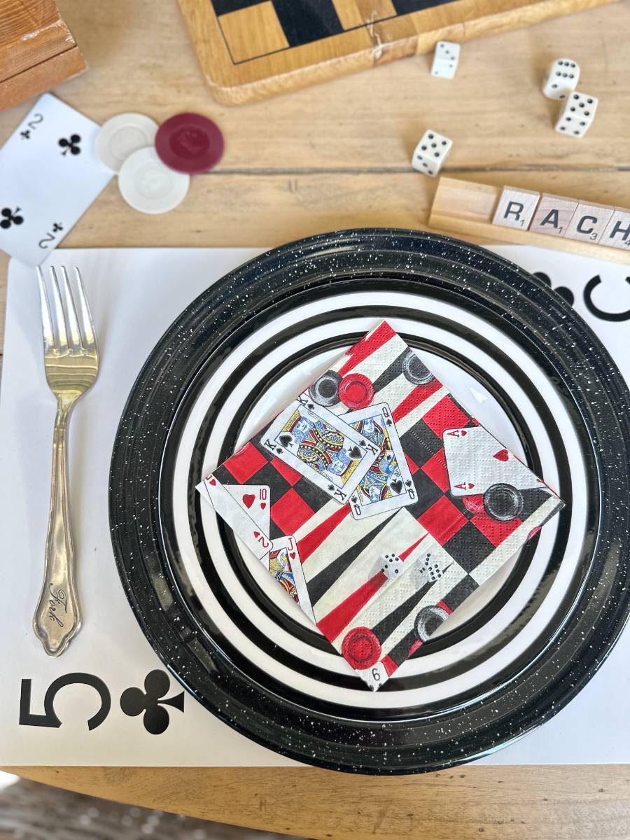 A table set for a game-themed meal features a black-and-white striped plate with a napkin depicting playing cards and checkers pieces. A fork rests to the left of the plate. Dice, game chips, a Scrabble letter holder, and a partial chessboard are visible in the background.