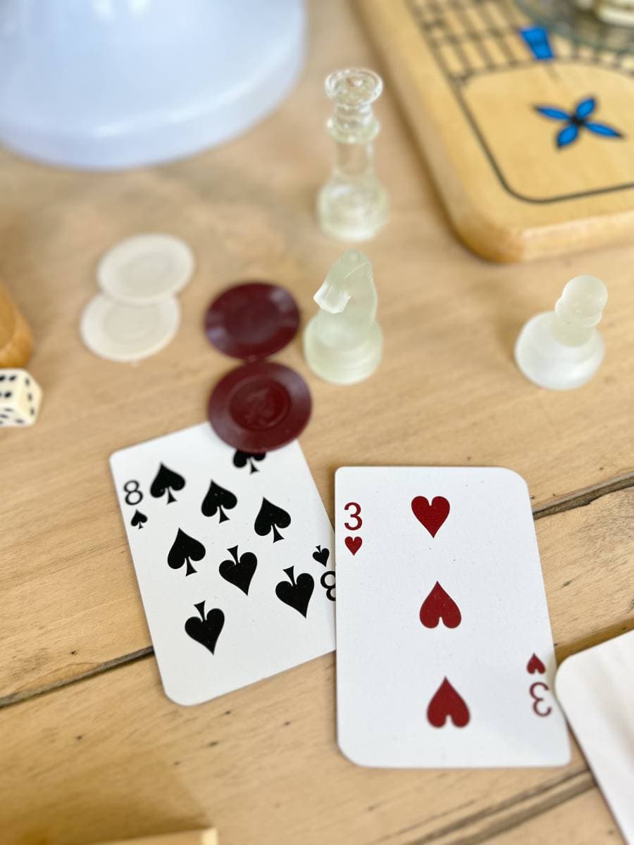 Two playing cards, an 8 of spades and a 3 of hearts, lie on a wooden table surrounded by game pieces including chess pieces, red and white checkers, and a die. A wooden game board with blue and black markings can be seen in the background.