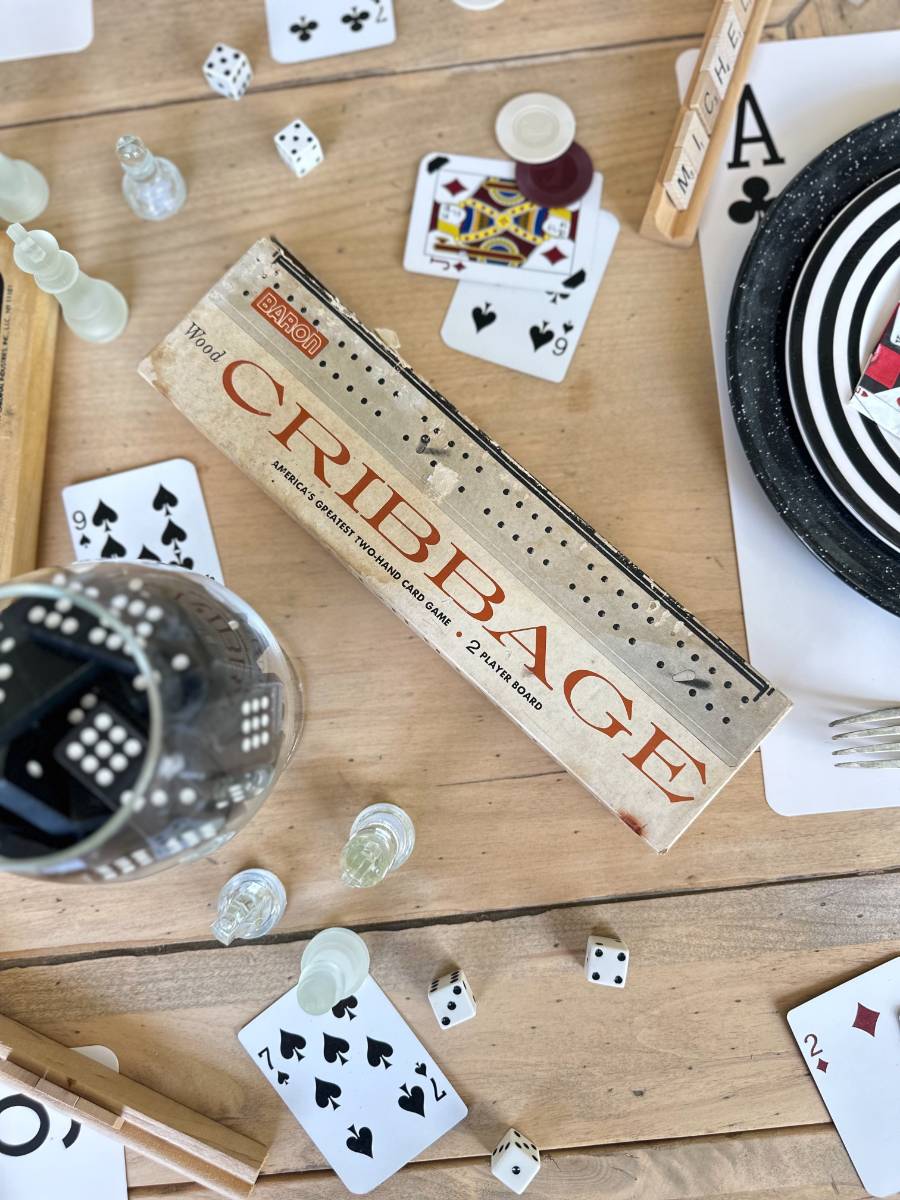 A wooden table is strewn with various game pieces including playing cards, dice, chess pieces, and a cribbage board game box. The cards and dice are scattered around casually, creating a playful and lively atmosphere. The focus is on the cribbage board game box in the center.