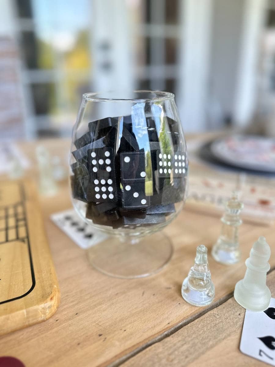 A glass container filled with black dominoes with white dots sits on a wooden table surrounded by various game pieces, including chess pieces and playing cards. A board game is partially visible in the background.