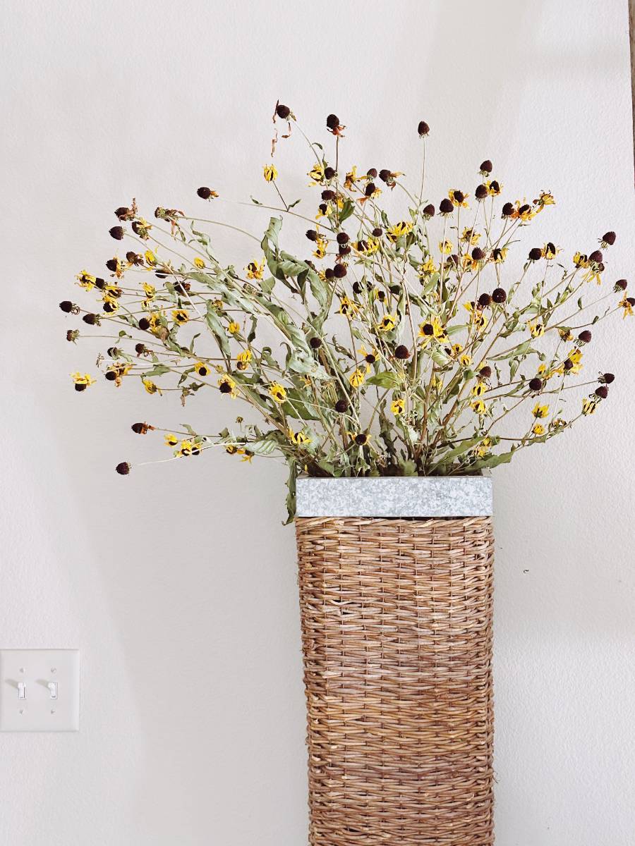 A woven wicker vase holds a bouquet of wilted yellow flowers with dark centers, capturing the change from summer to fall on a budget, set against a plain white wall with a light switch visible in the lower left corner.