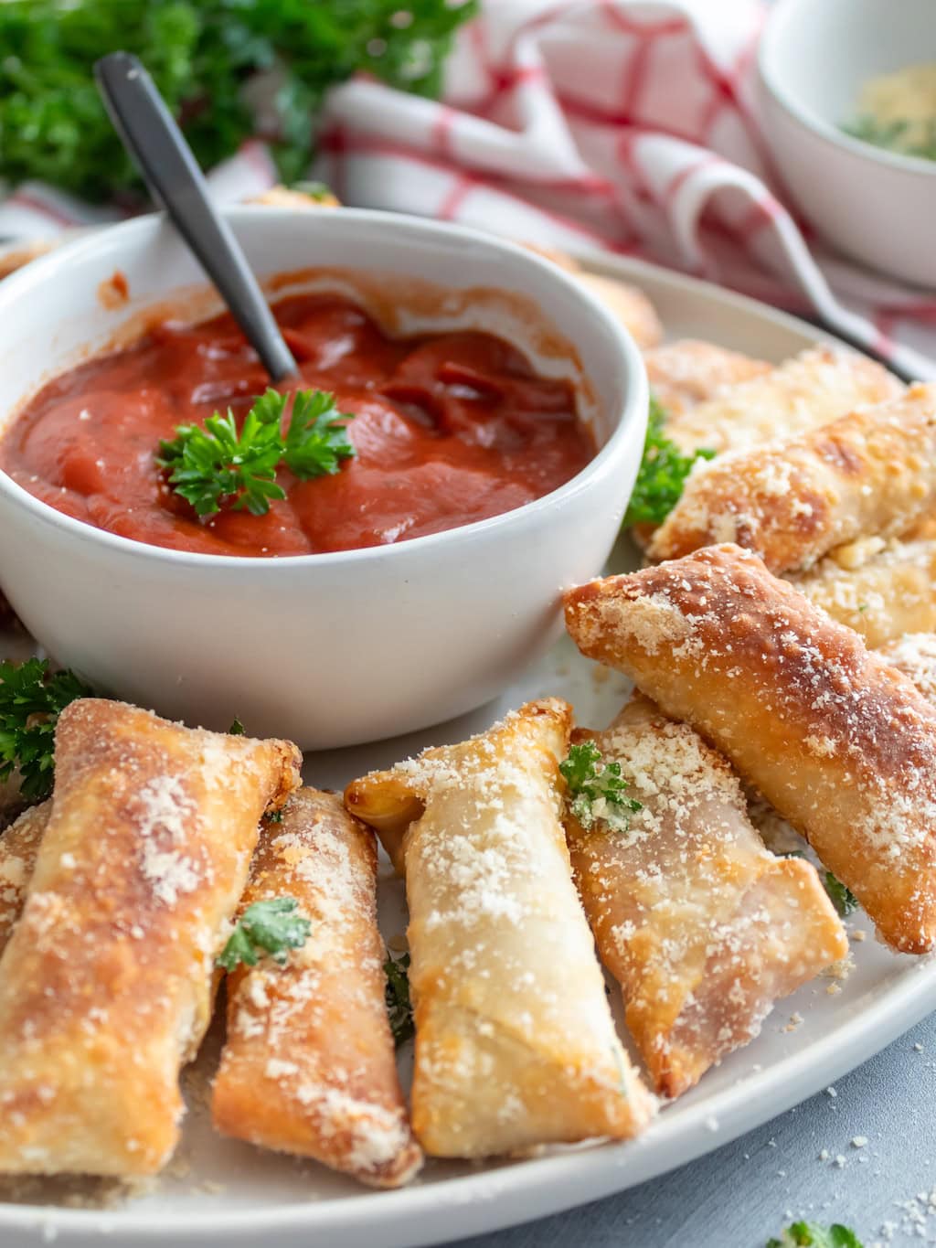 A white plate filled with crispy, golden-brown pastry rolls dusted with grated cheese is set beside a small white bowl of red dipping sauce. A spoon is in the sauce, and fresh parsley garnishes both the plate and bowl. Perfect for recipes for a Winning Game Night Party, a checked cloth lies in the background.