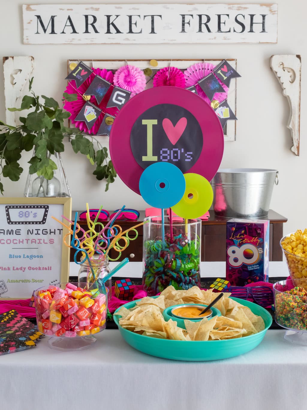 A colorful 80s-themed party table setup features a sign that reads "I ♥ 80's", neon decorations, a plate of chips, bowls of candy, and a menu for 80's Game Night Cocktails. A "Market Fresh" sign is displayed above the table.